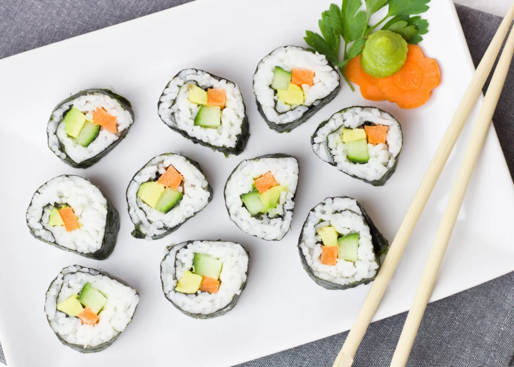 Veganes Sushi mit Avocado und Karotte auf einem eckigen Teller, Stäbchen auf dem Rand des Tellers